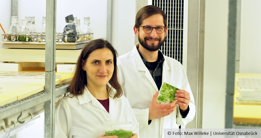 Two smiling people in lab coats holding boxes with little plants.