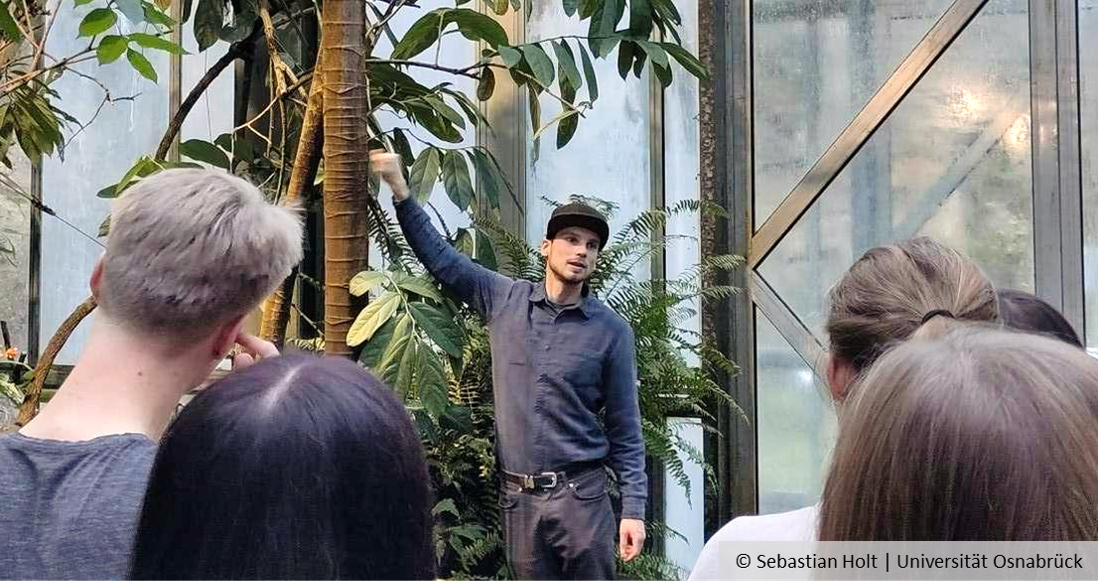 People stand in the rainforest house of the botanical garden and look at a man who is apparently explaining something.