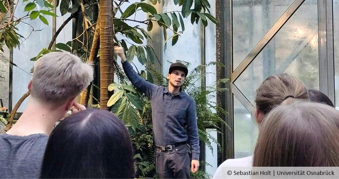 Personen stehen im Regenwaldhaus des Botanischen Gartens und schauen auf einen Mann, der offenbar etwas erklärt. Personen stehen im Regenwaldhaus des Botanischen Gartens und schauen auf einen Mann, der offenbar etwas erklärt.