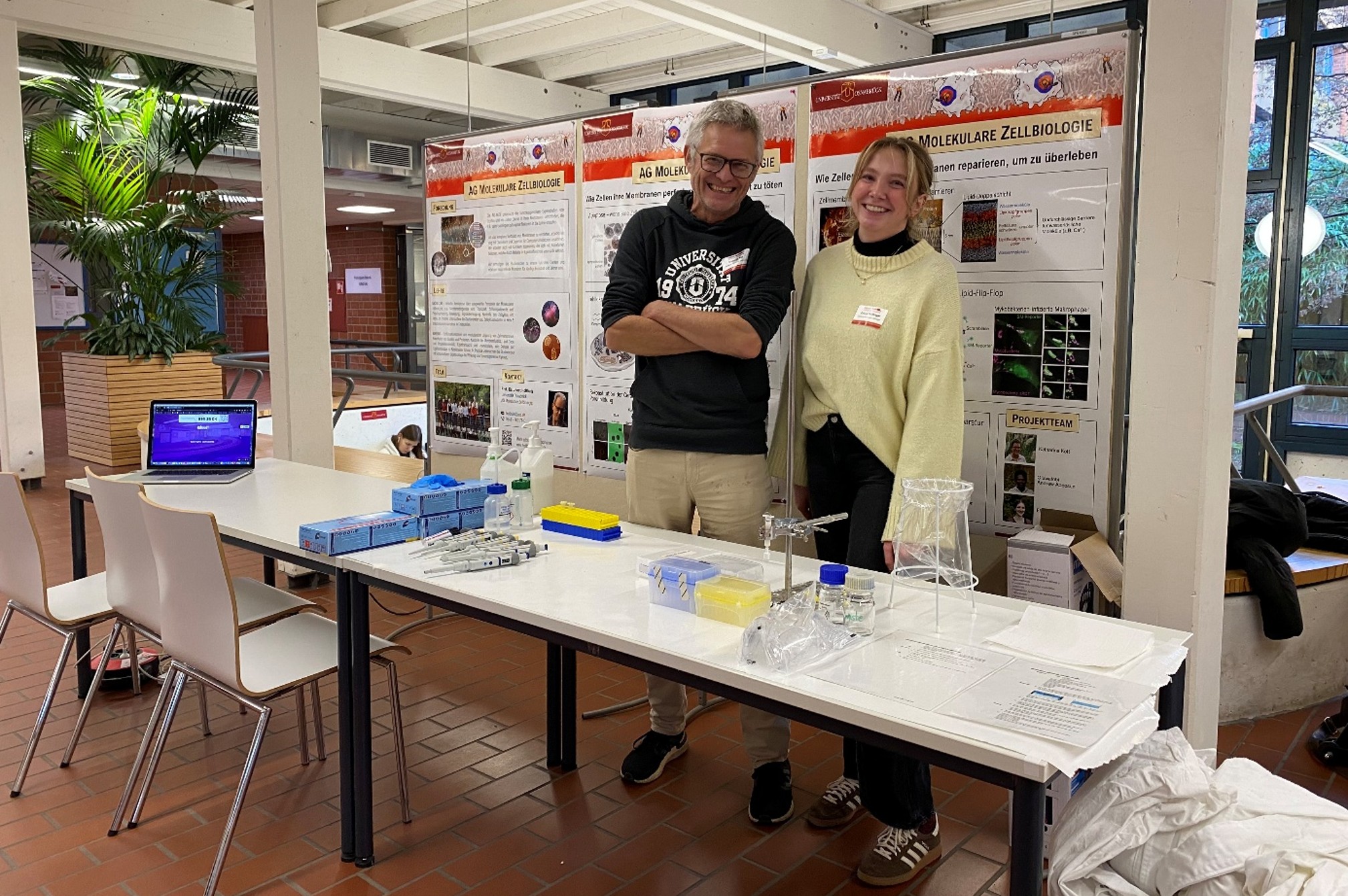 A man and a woman are standing behind a table with various laboratory items on it, smiling.