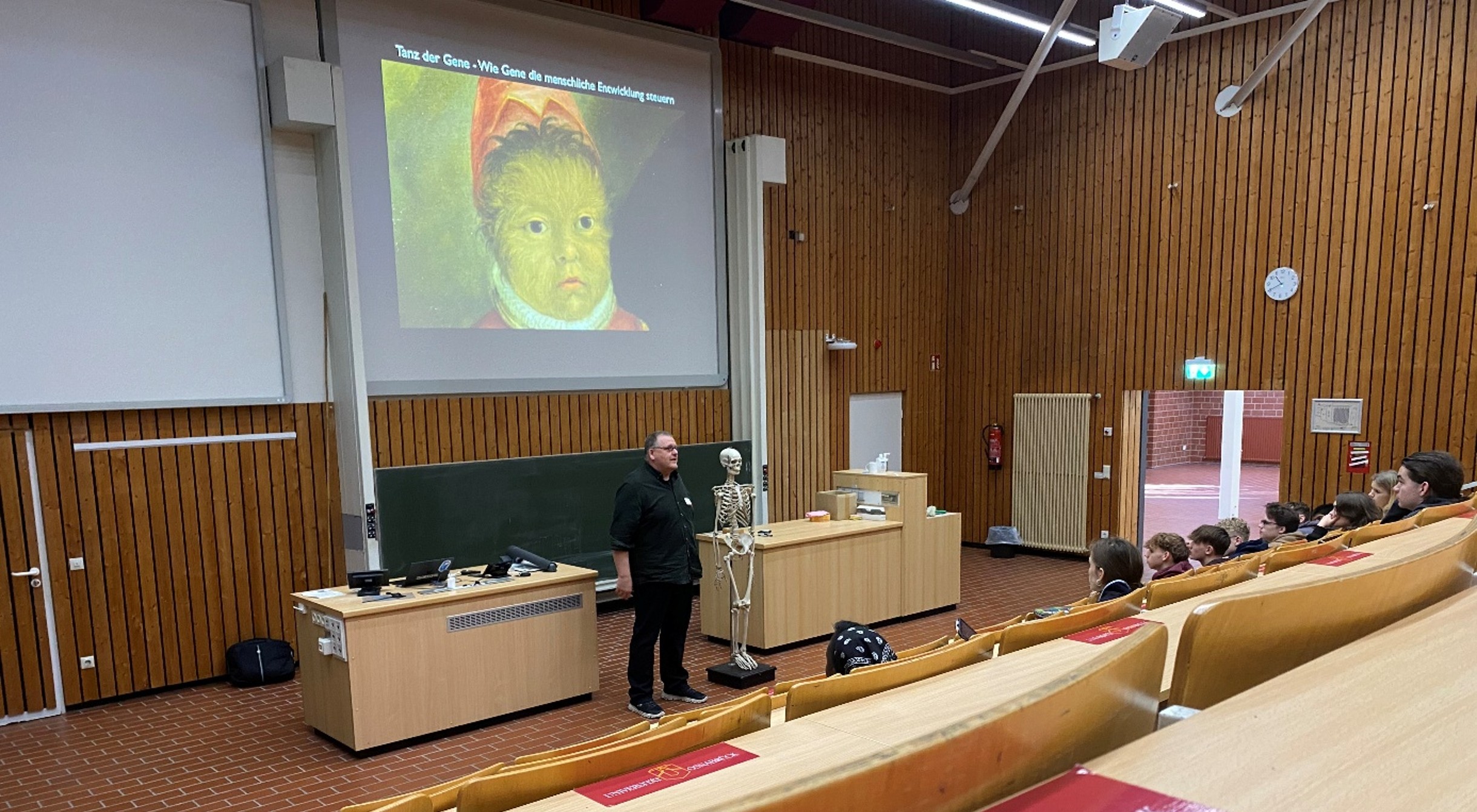 A man is standing in a lecture hall, next to him is a human skeleton.
