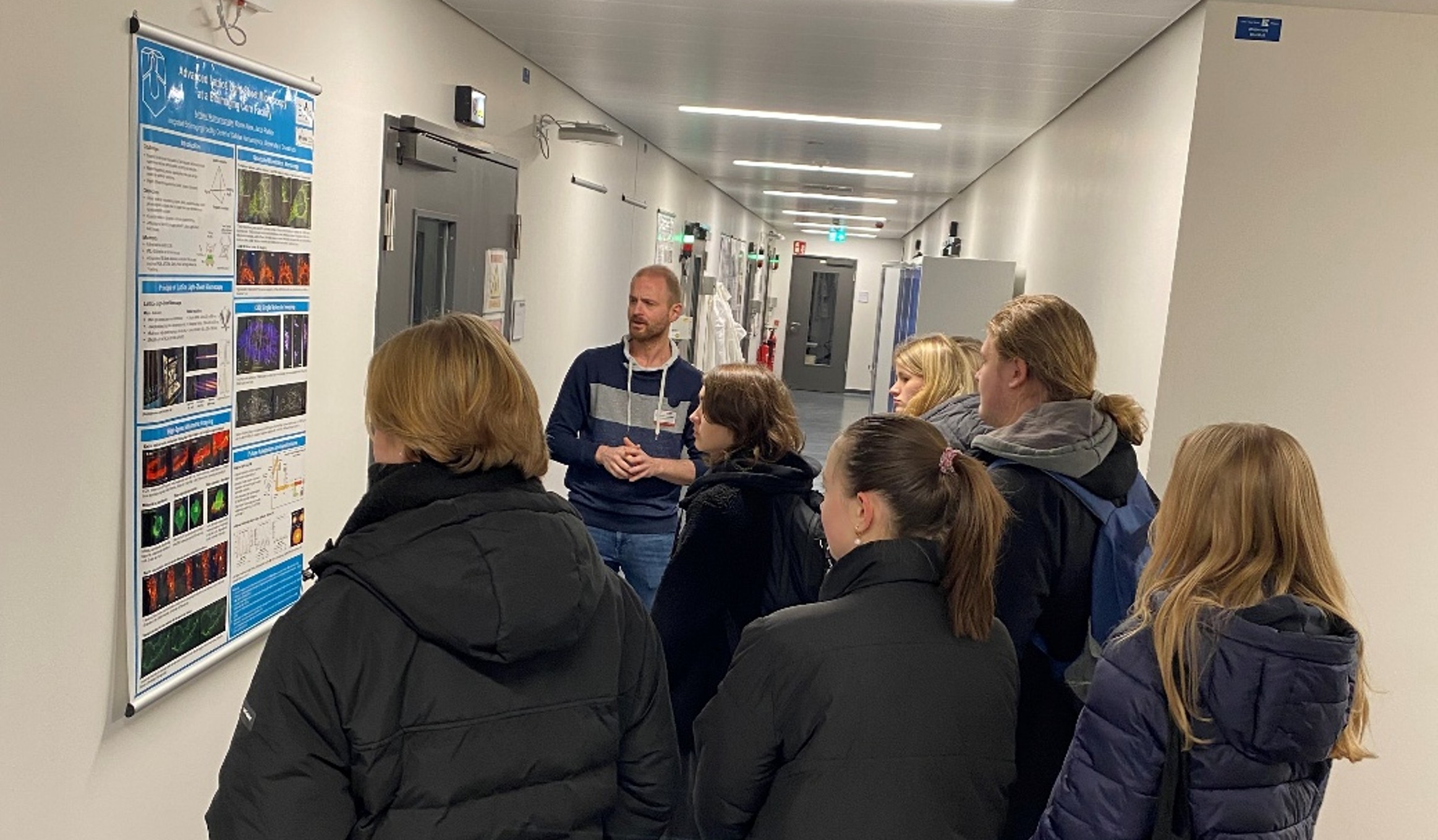 A man and a group of pupils are standing in a corridor in front of a poster.