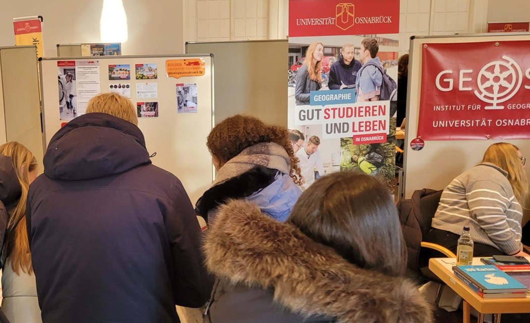 Pupils stand crowded together, with posters and information material bearing the Osnabrück University logo visible in the background.