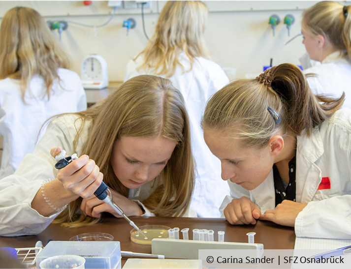 Zwei Schülerinnen schauen auf eine Petrischale, eine hantiert mit einer Pipette.